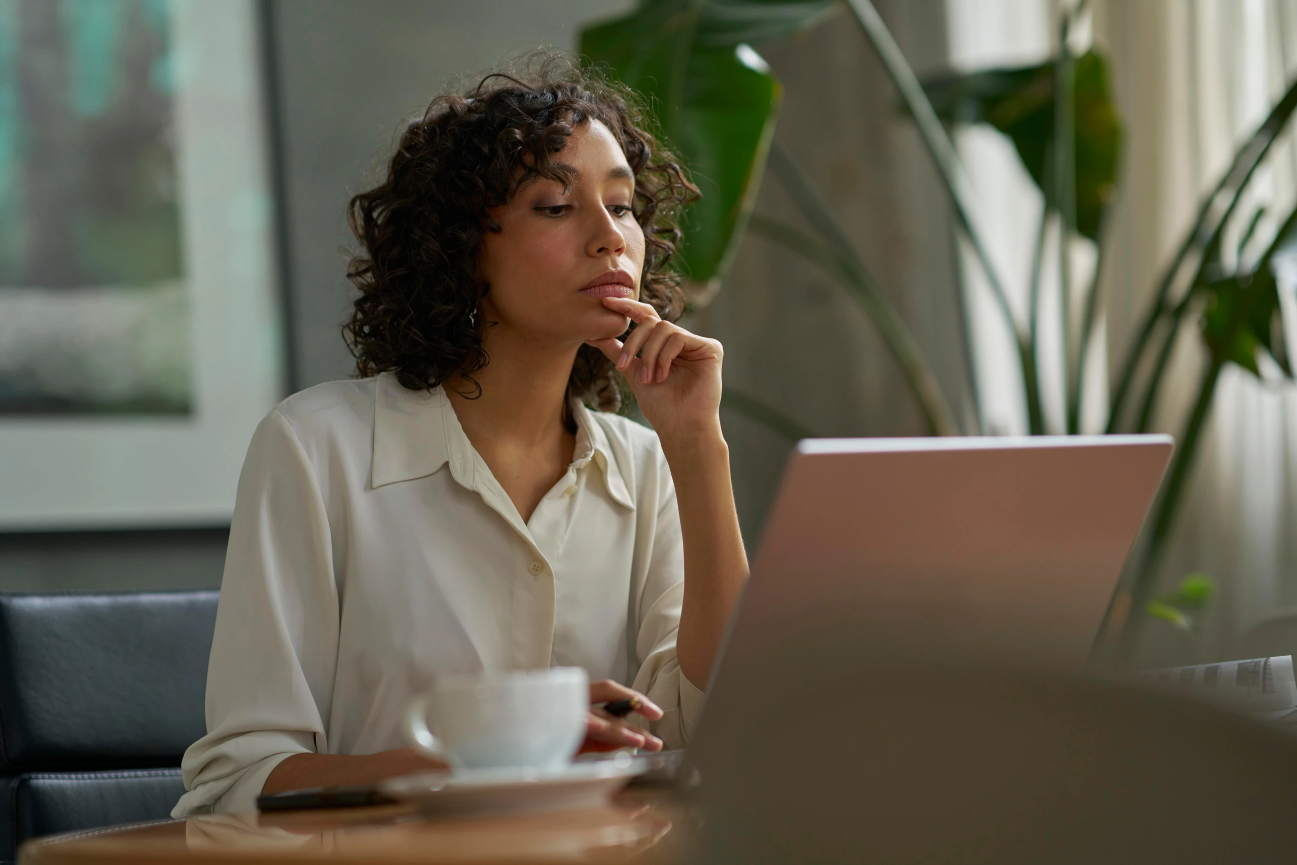 A woman with curly hair wearing a white shirt sits at a desk, looking thoughtfully at her laptop screen with her hand resting near her chin.
