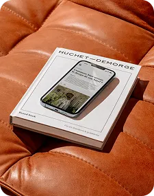Un livre blanc titré Huchet-Demorge avec un smartphone affichant un site sur un canapé en cuir marron.