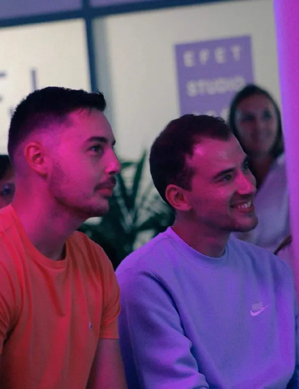 Deux hommes assis regardant attentivement vers la droite sous un éclairage violet et rose.