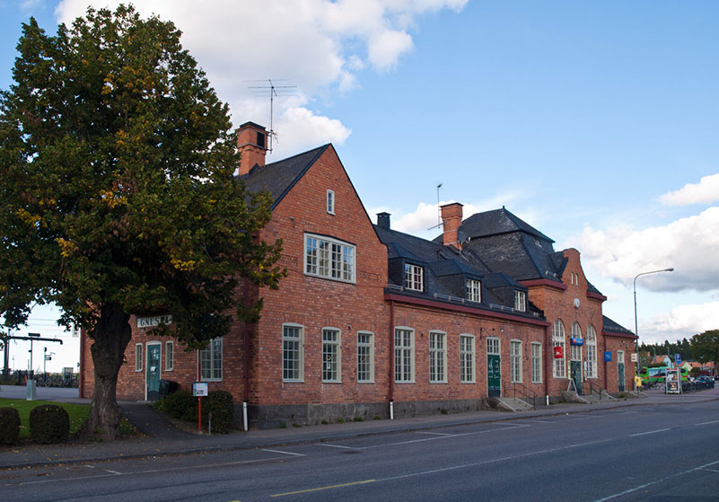 Gnesta stationsbyggnad i rött tegel och svart plåttak med träd i förgrunden.