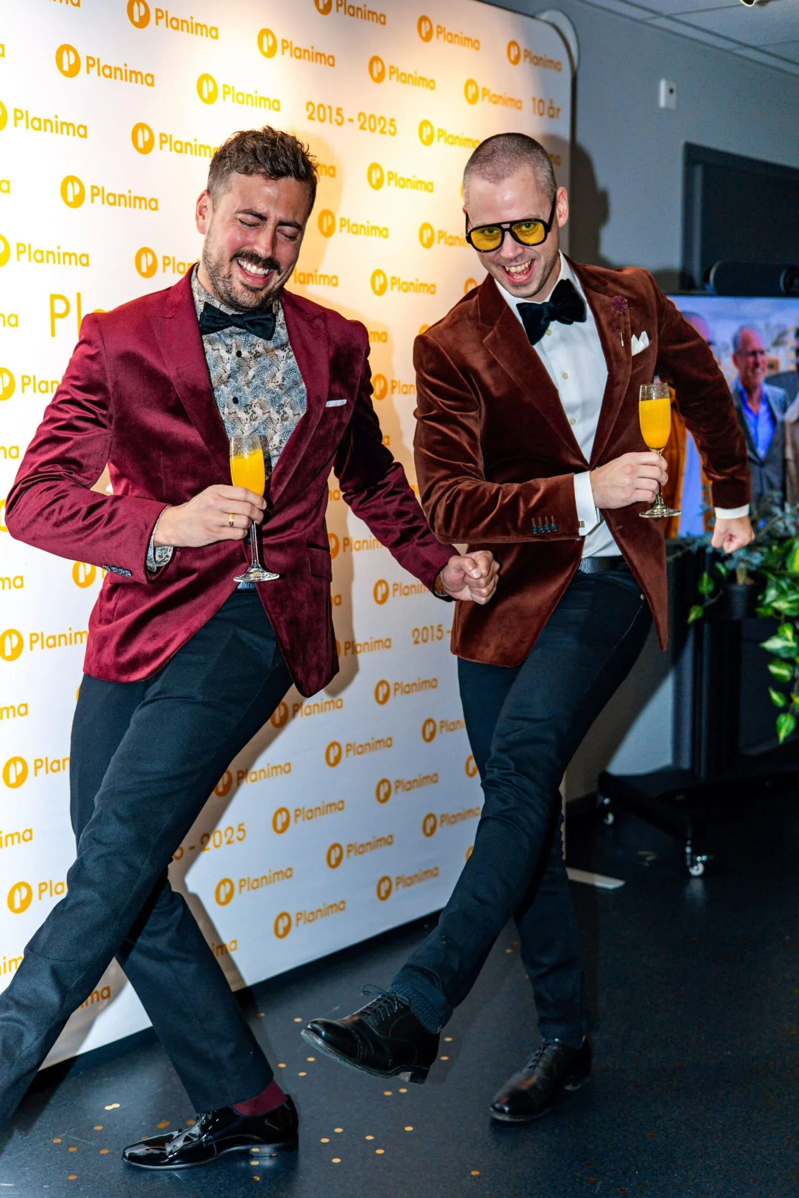 Jens och Anton dansar i sammetskavajer framför Planimas 10-årsbakdrop på jubileumsfesten