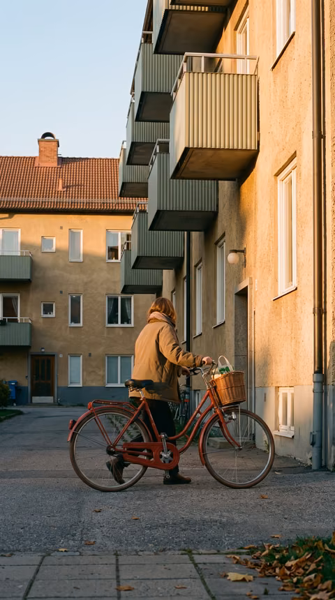 Person som leder en röd cykel med korg över en innergård mellan flerbostadshus med gröna balkonger i kvällssol