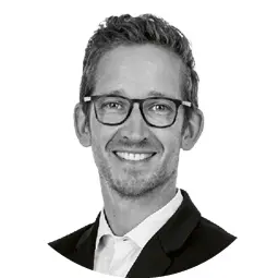 Porträt eines Mannes mittleren Alters mit kurzen welligen Haaren und Brille, der in einem weißen Hemd und dunklem Sakko lächelnd in die Kamera blickt.