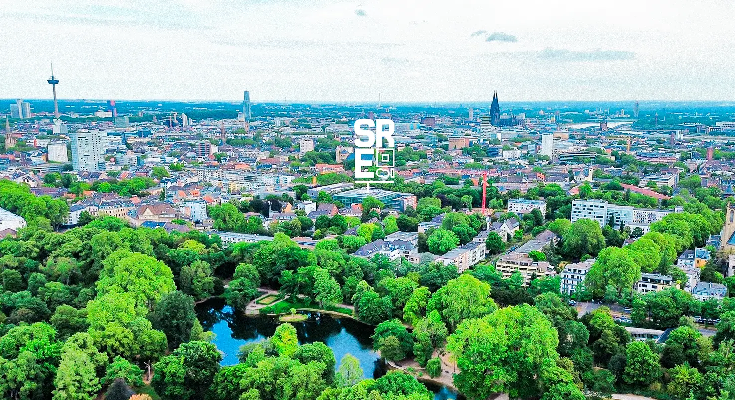 Luftaufnahme von Köln mit grünen Bäumen und Teichen im Volksgarten im Vordergrund, Wohngebieten dahinter, und dem Kölner Dom sowie Fernsehturm am Horizont.