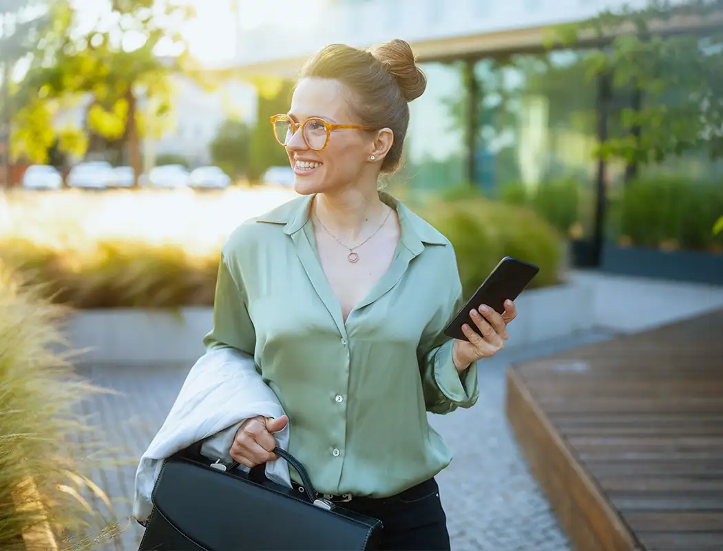 Frau mit zusammengebundenem Haar, trägt Brille und grüne Bluse, hält Smartphone in der rechten Hand und schwarze Aktentasche in der linken Hand, geht draußen auf einem Weg mit moderner Architektur im Hintergrund.