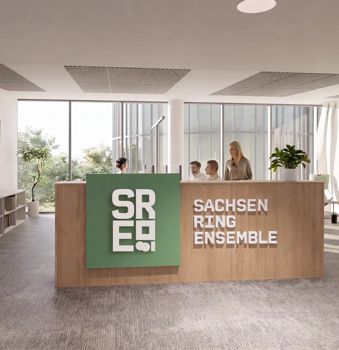 Moderner Empfangsbereich mit hölzerner Theke, auf der "Sachsen Ring Ensemble" und ein grünes Logo mit den Buchstaben S, R, E und zwei Punkten stehen, im Hintergrund drei sitzende und eine stehende Person, große Fenster und Pflanzen.