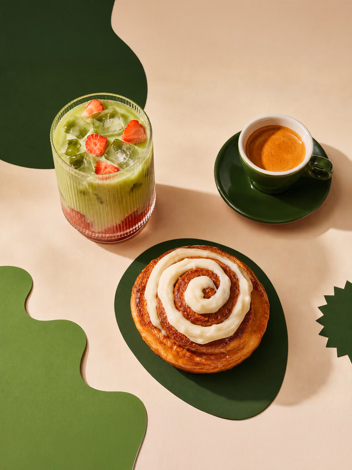 Iced green matcha drink with strawberries, espresso in green cup, and cinnamon roll with icing on green shapes.
