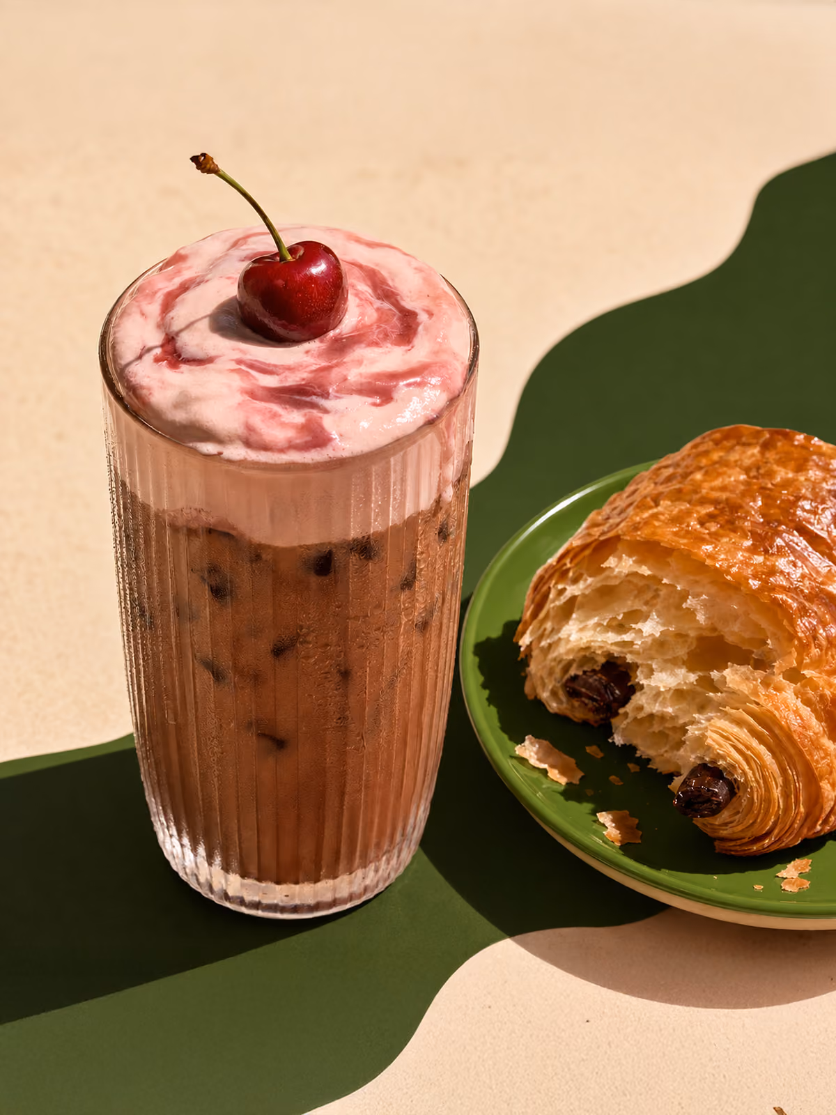 Glass of iced chocolate drink topped with pink whipped cream and a cherry, beside a chocolate croissant on a green plate.