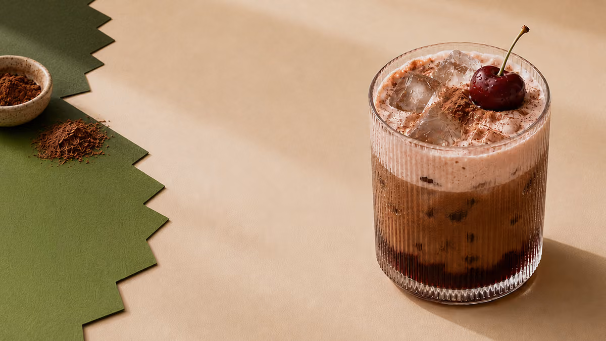 Iced chocolate drink with foam, ice cubes, grated chocolate, and a cherry in a ribbed glass on beige surface.