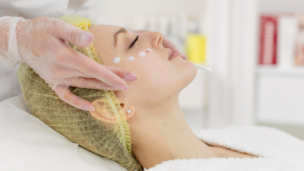 Woman with closed eyes lying down receiving facial cream treatment in a spa or clinic.