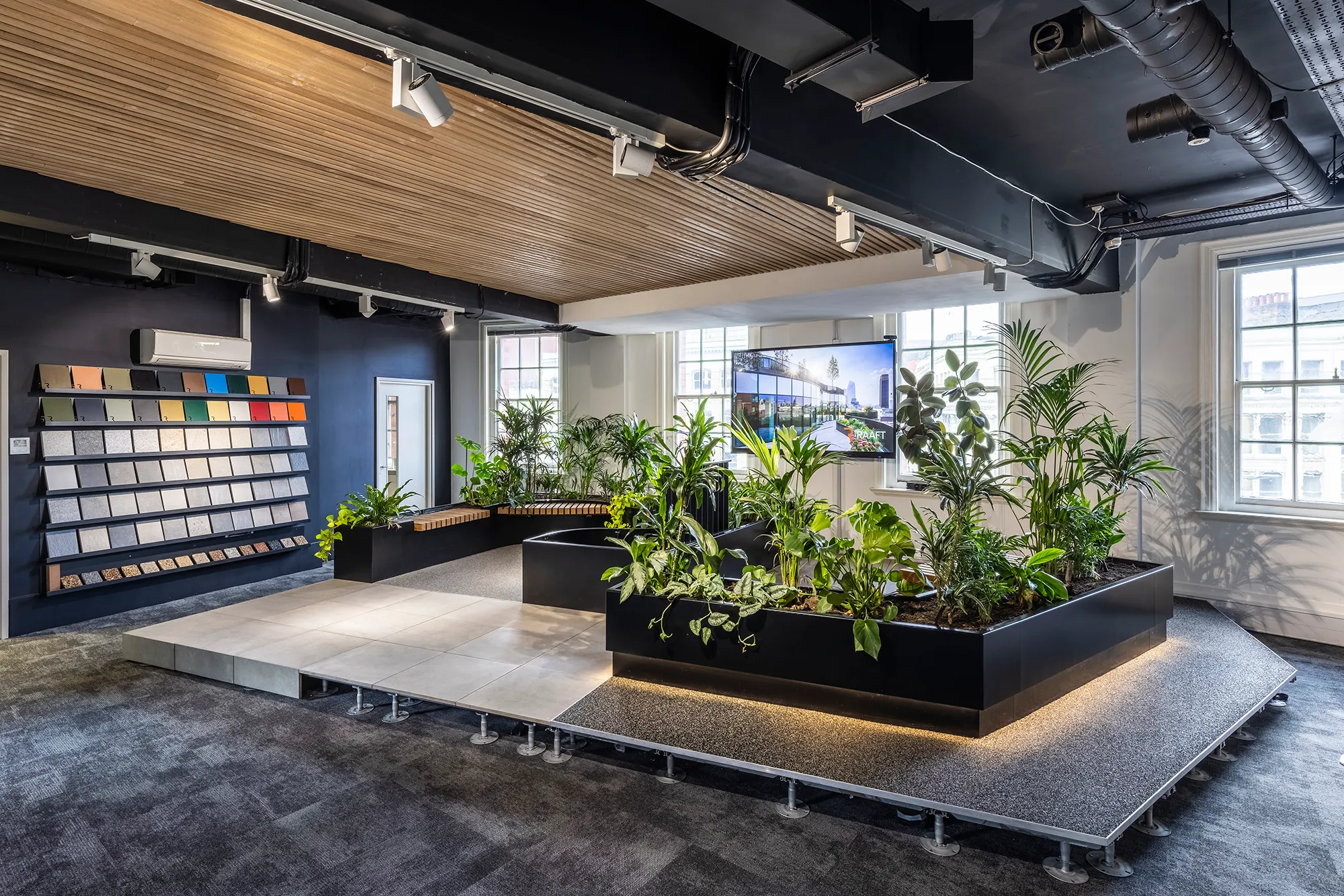 Modern showroom with samples on wall, central platform with plants, seating, and a large screen by windows.