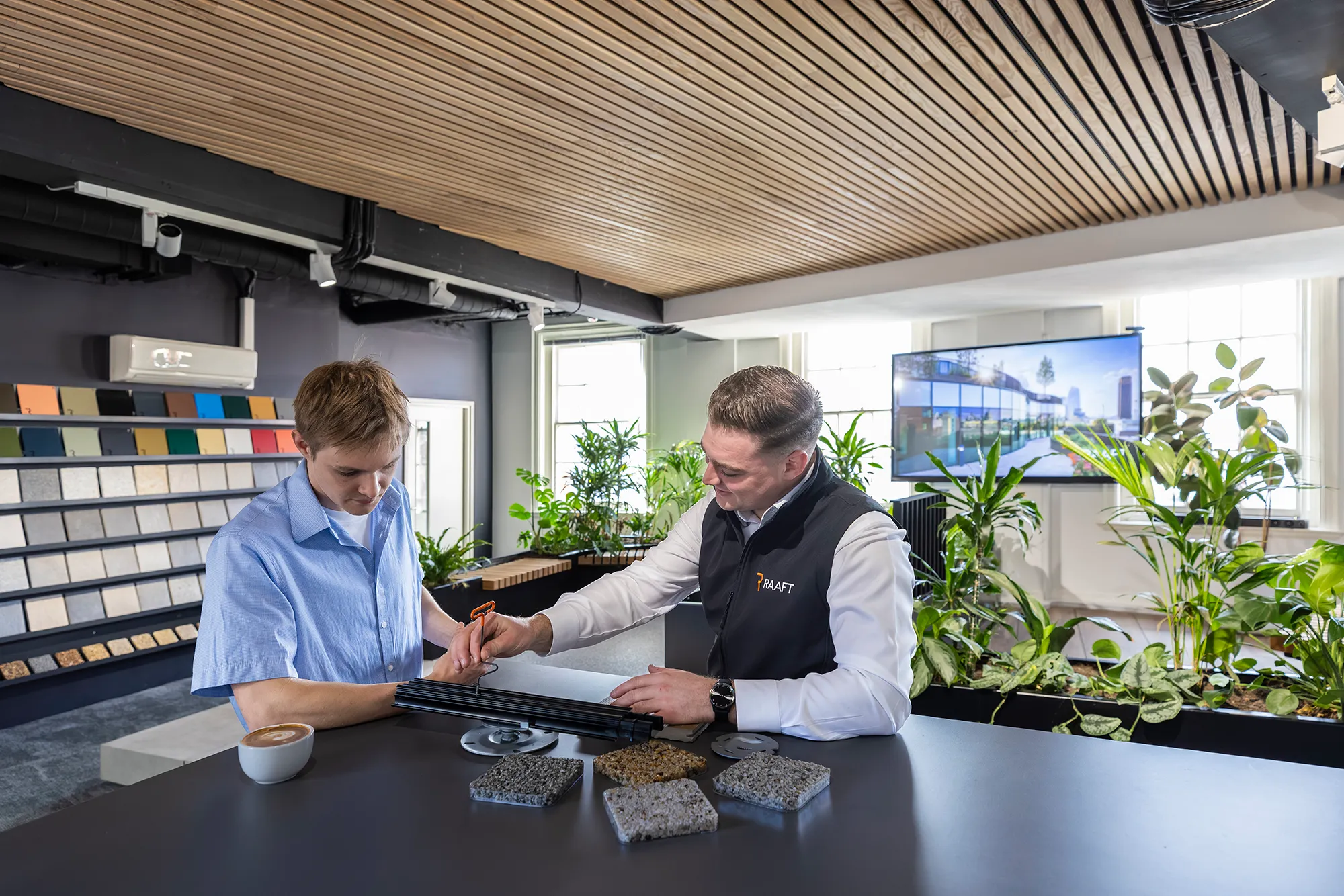 Two men discussing black samples at a table with stone swatches and plant-filled showroom background.