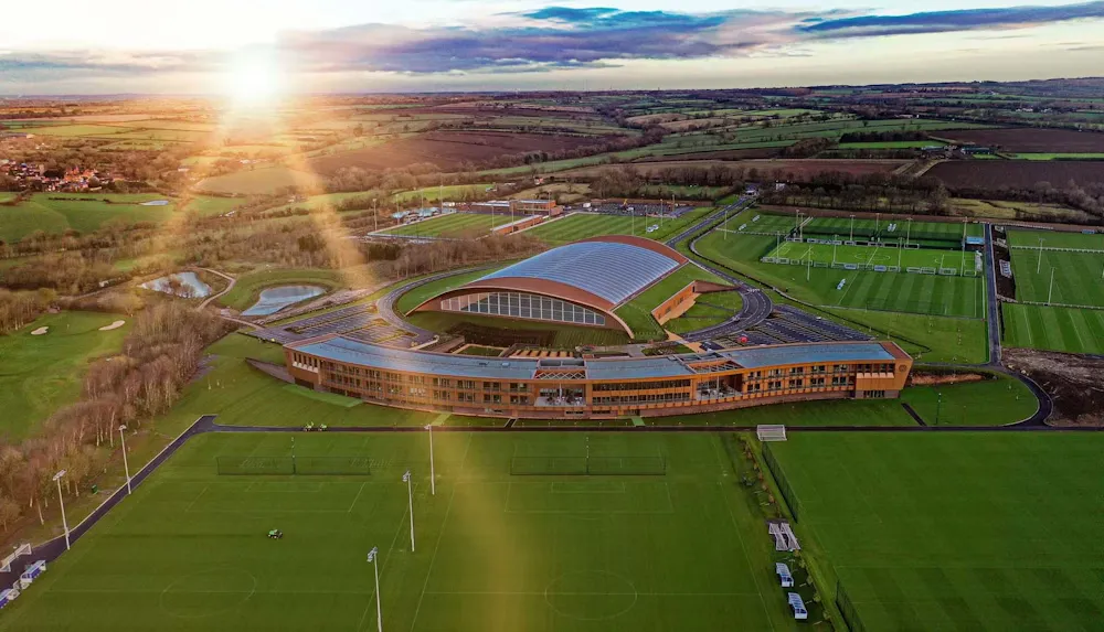 Aerial view of a sports training ground with green fields, a large curved building, and sunlight over the landscape.