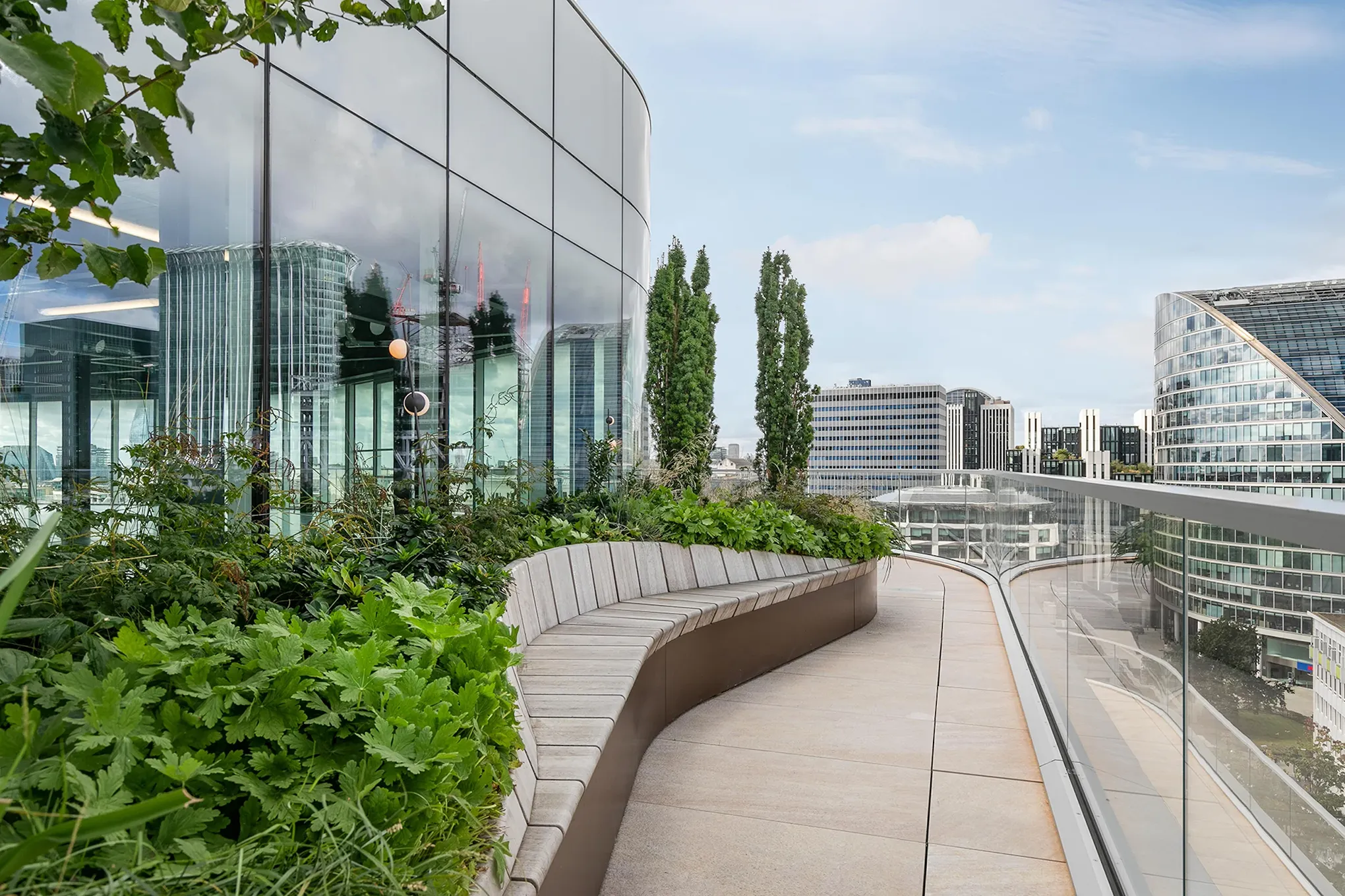 Modern rooftop terrace with curved bench, greenery, glass railing, and city buildings in background.