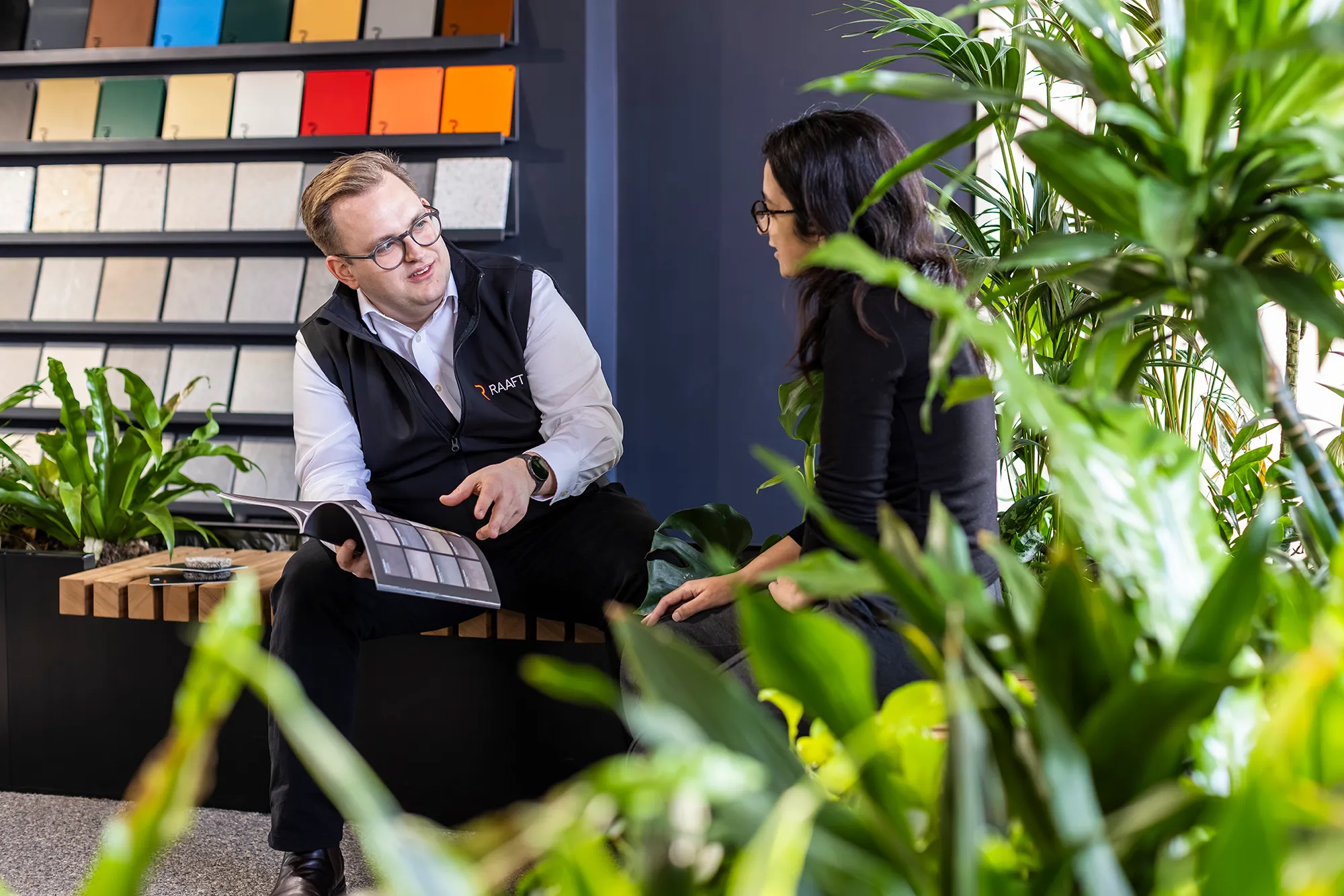 Man in glasses and vest shows design samples from a book to a woman seated among green plants.