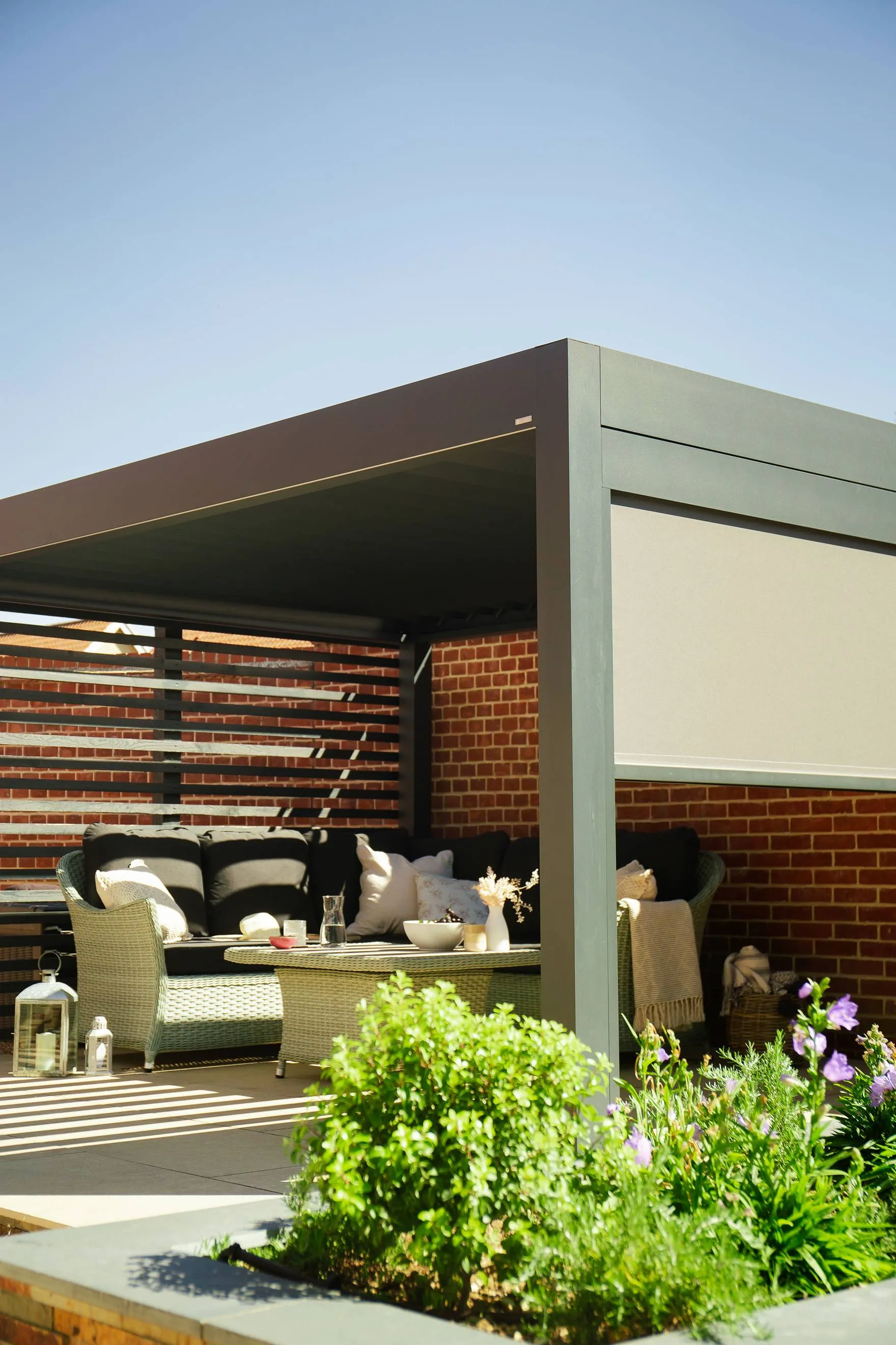 Modern outdoor pergola with wicker seating, cushions, coffee table, and garden plants in front.