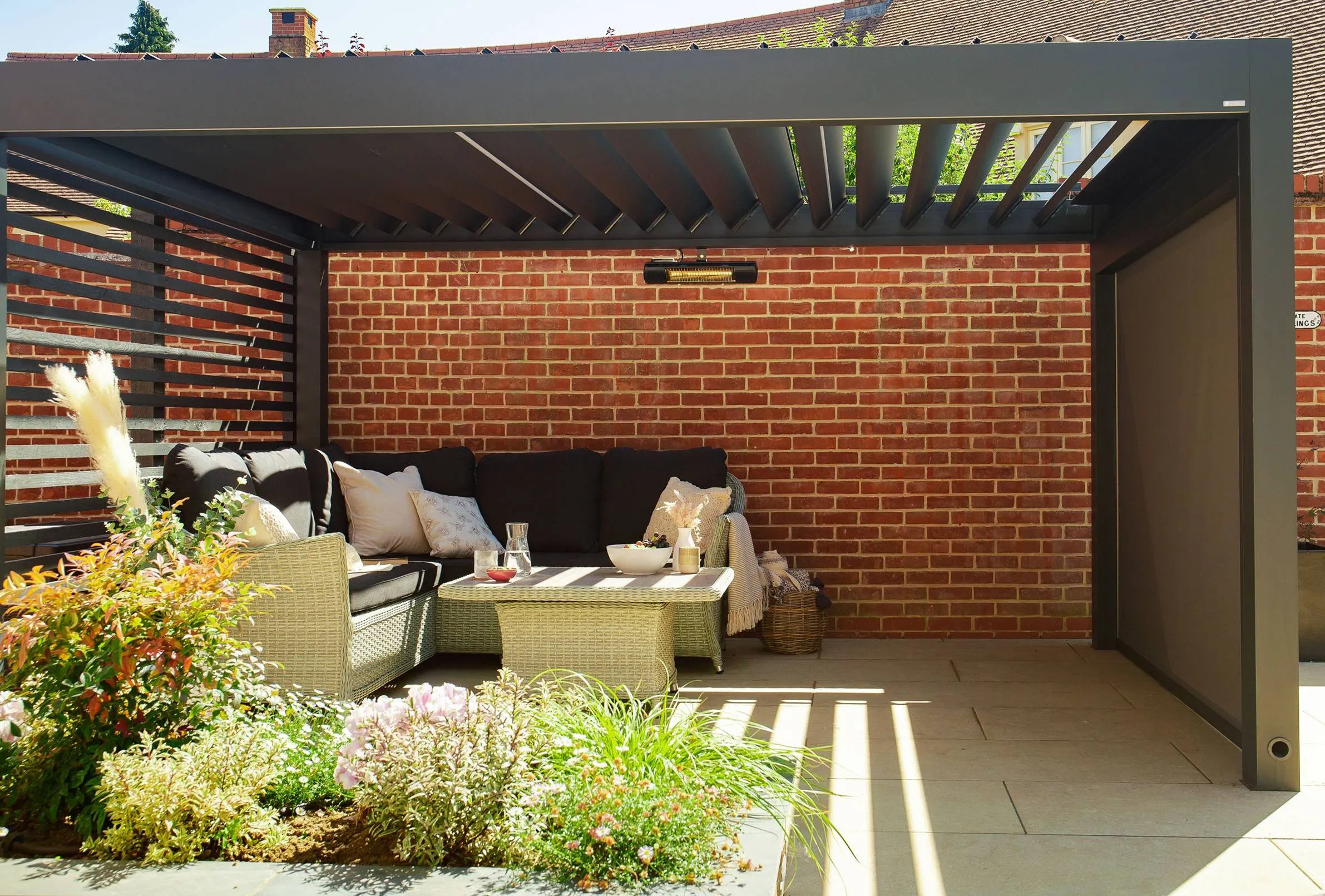 Modern outdoor pergola with black sectional sofa, beige cushions, coffee table, and plants in front.
