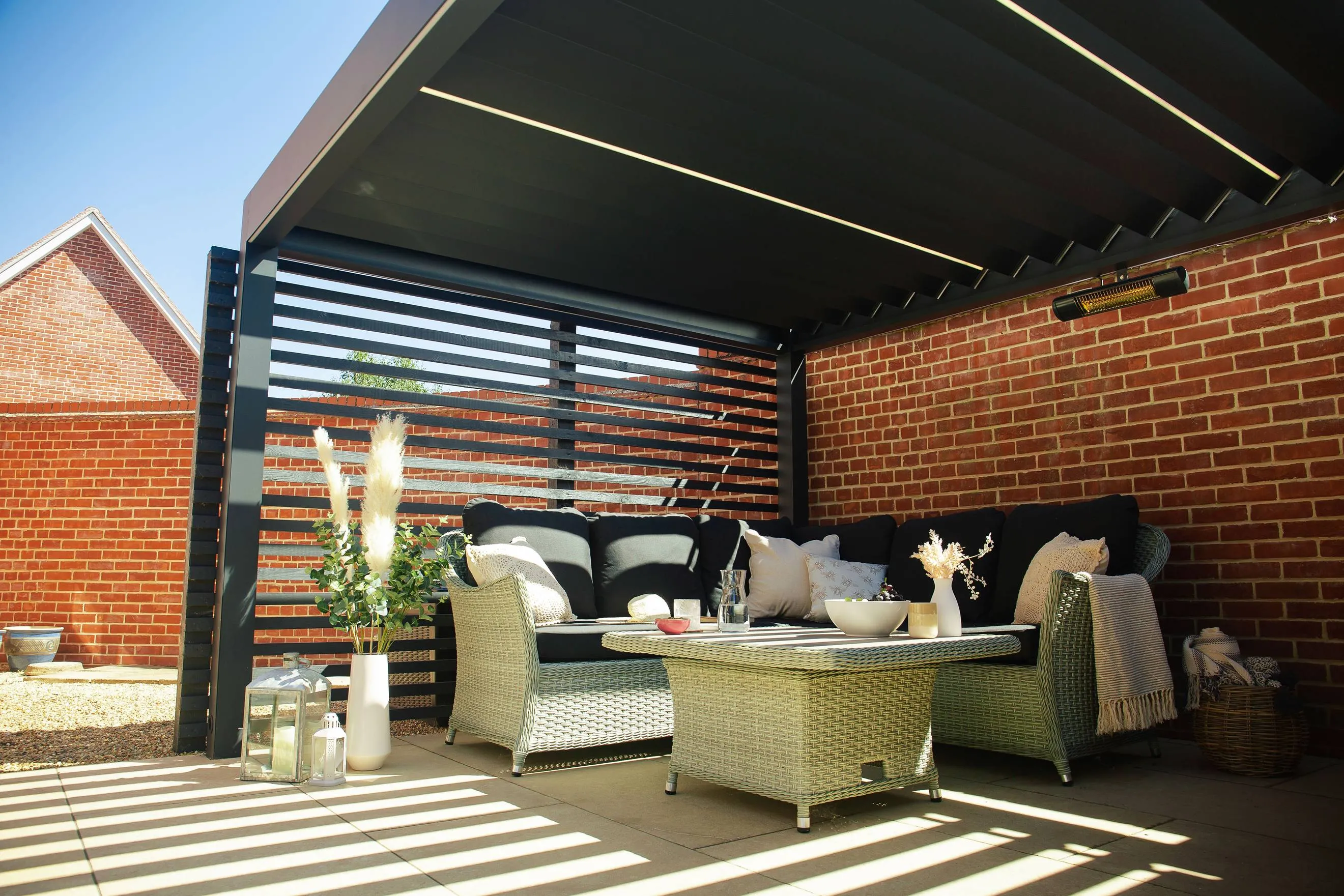 Outdoor patio seating with wicker sofa, black cushions, pillows, and decorative table under shaded pergola.