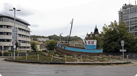 Small blue and brown boat displayed on grass in urban roundabout with buildings and trees around.