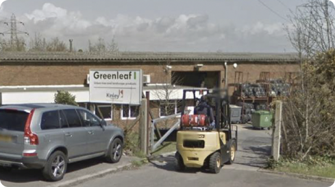 Forklift carrying items entering a Greenleaf garden products warehouse with a parked silver SUV outside.