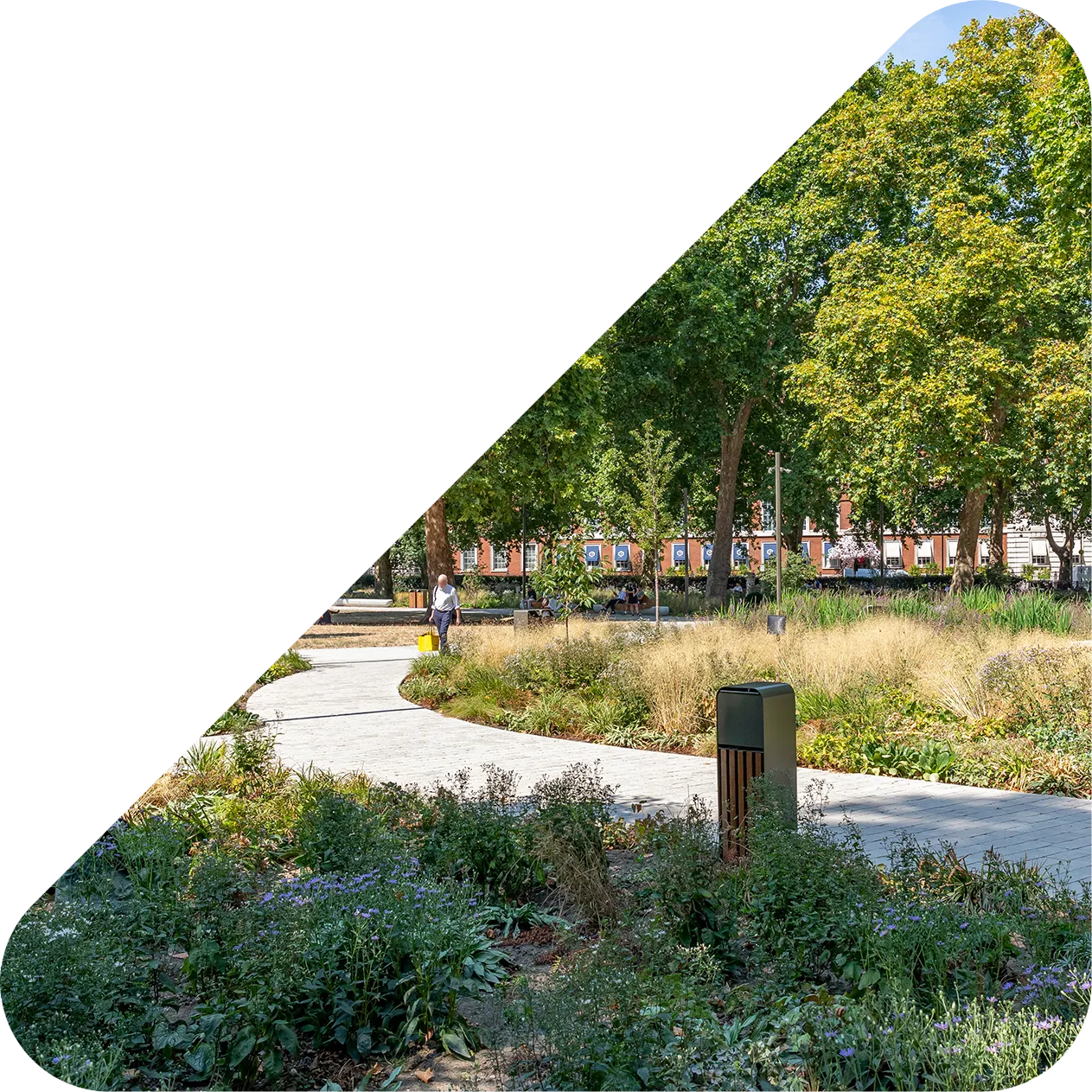 Curved walkway in a green park with trees, plants, and a person walking carrying a yellow bag.