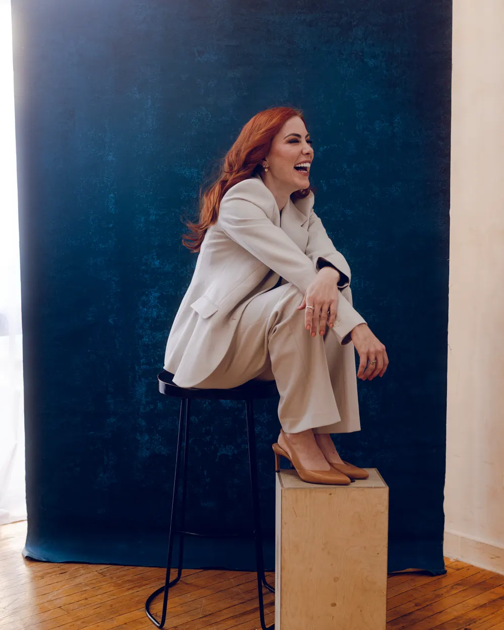 Katharine Quinn laughing, seated on a stool in a cream suit in front of a deep blue painted studio backdrop