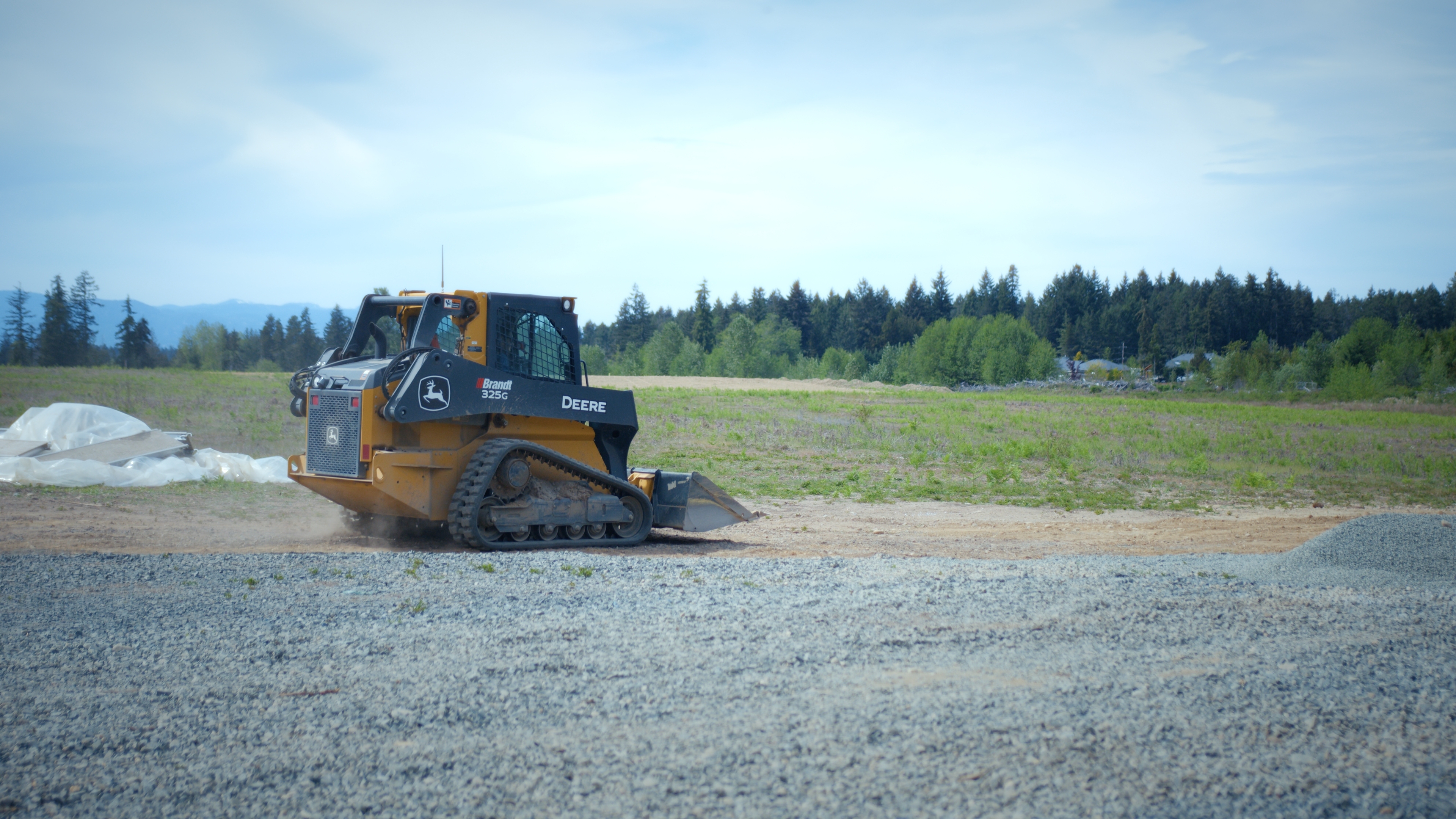 John Deere 325G skid steer loader on gravel near a field with trees under a blue sky.
