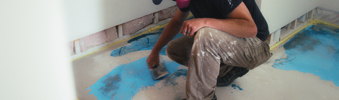 Person wearing a respirator mask applying blue waterproofing on a floor with a trowel.