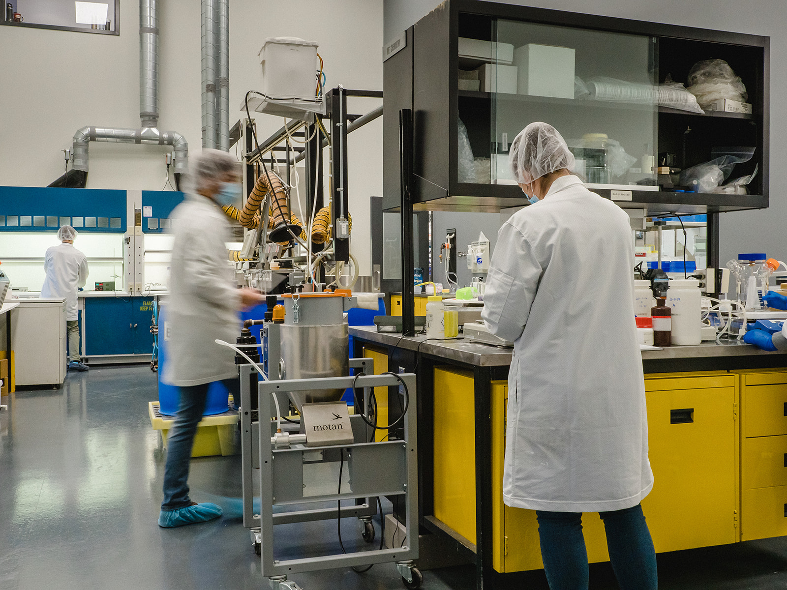 A laboratory scene with three people wearing white lab coats, hair nets, masks, and shoe covers working among yellow cabinets and scientific equipment.