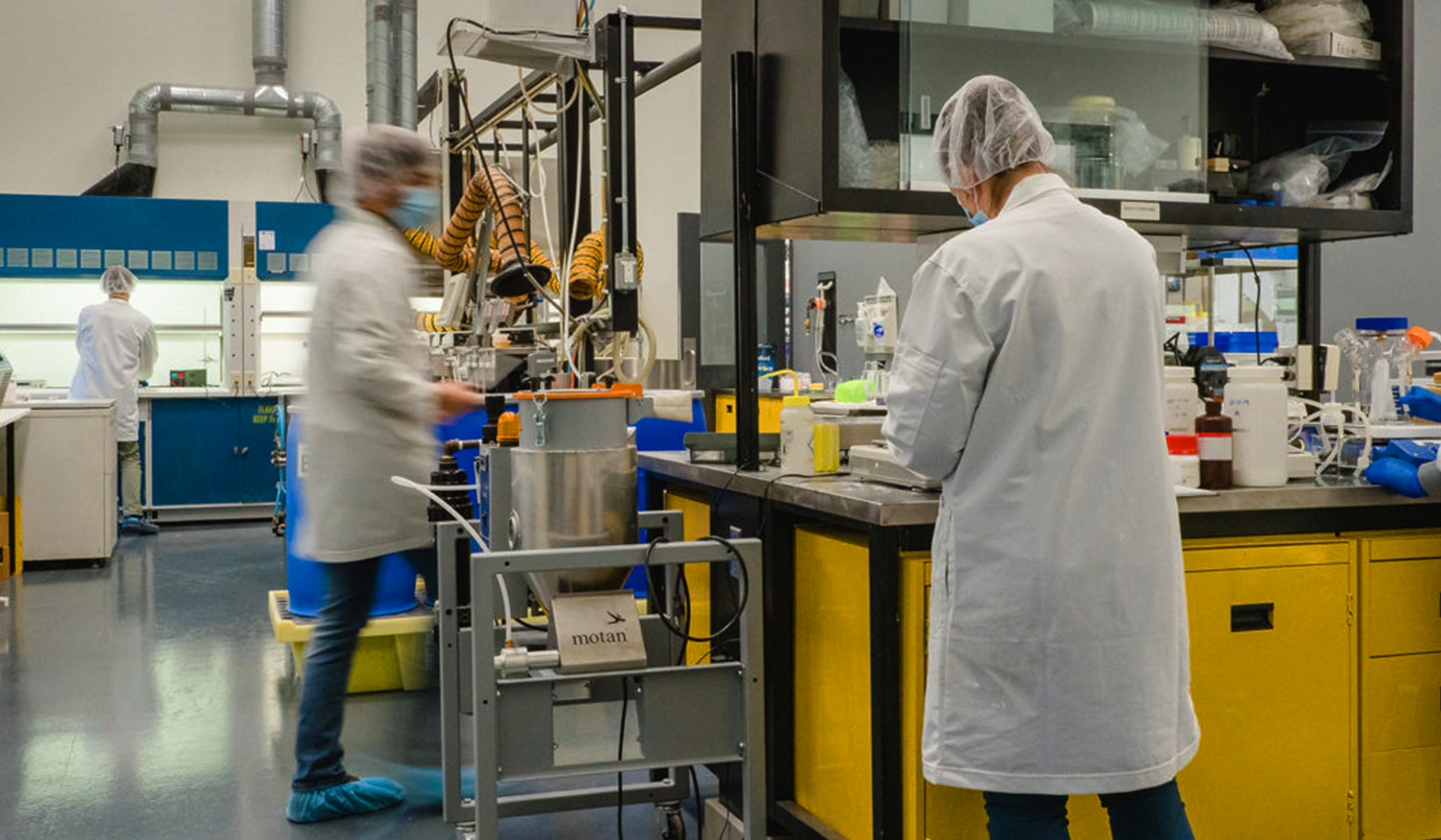 Researchers wearing white lab coats, hairnets, masks, and shoe covers working in a laboratory with scientific equipment, yellow cabinets, and blue ventilation ducts.