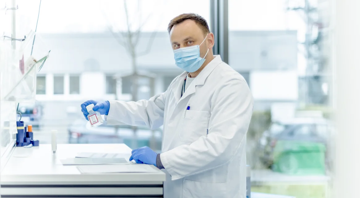 Scientist wearing a lab coat, mask and blue gloves holds a sample bottle at a laboratory bench.