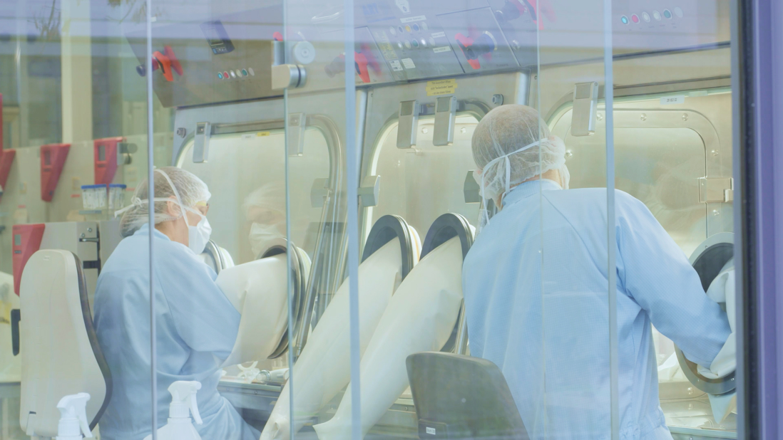 Two laboratory workers in protective clothing and hairnets operate isolator glove boxes in a cleanroom setting.