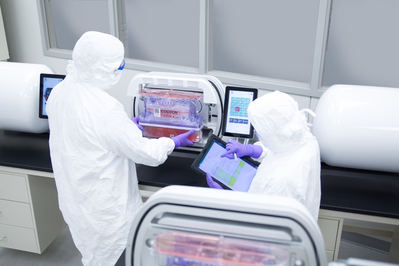 Two operators in cleanroom suits working with a Cocoon Platform instrument, one handling the unit and one using a tablet.