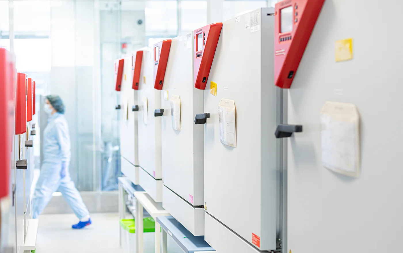 Row of white laboratory incubators with red control panels on top, with a person in full protective gear walking in the background inside a bright, clean lab.