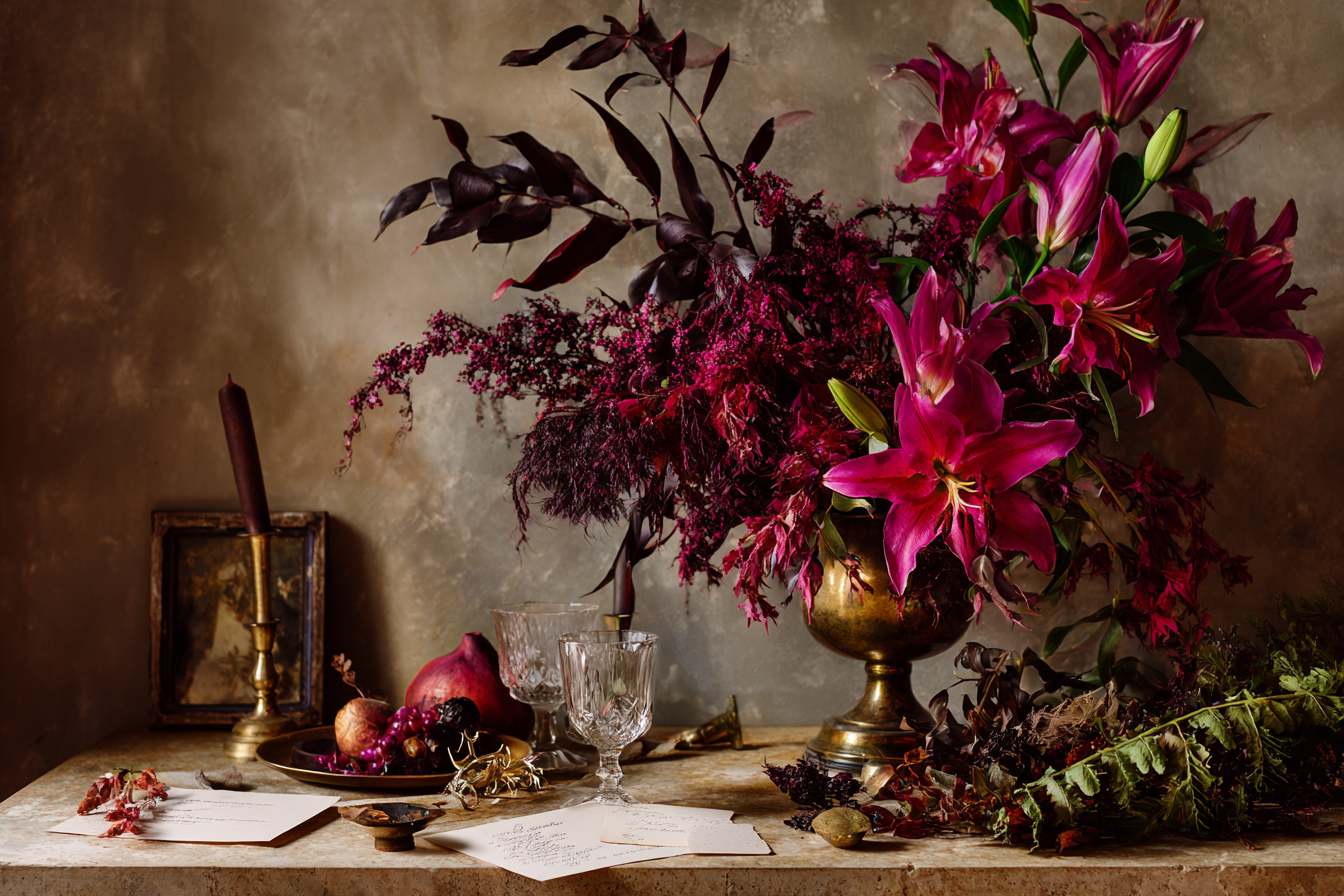 Vintage still life with dark red flowers, brass vase, candle, fruit, glasses, and handwritten letters on table.