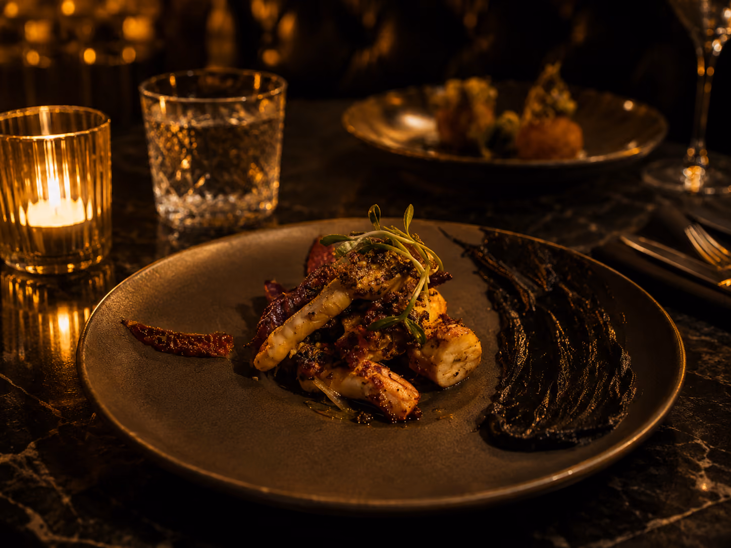Gourmet dish with grilled seafood, herbs, and dark sauce on a brown plate in candlelight.