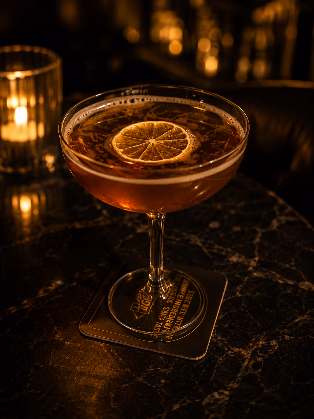 Cocktail in a coupe glass with a lemon slice garnish on a dark marble table with candlelight.