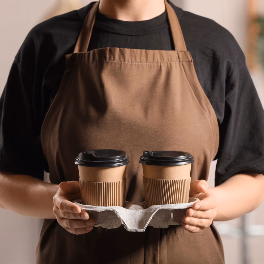A woman wearing a brown apron holding two coffee cups.