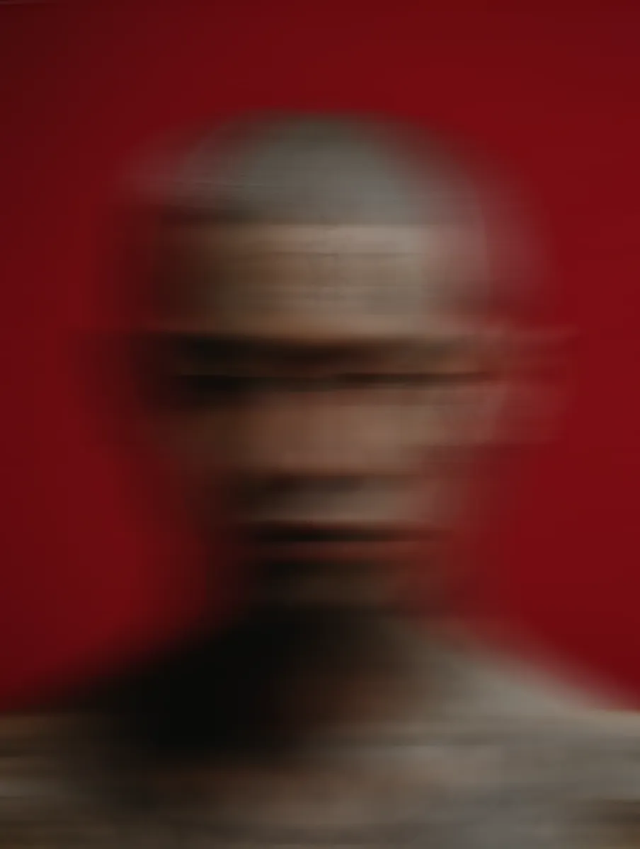 A heavily blurred and distorted portrait of a bald person against a red background, creating an abstract and unclear facial image.