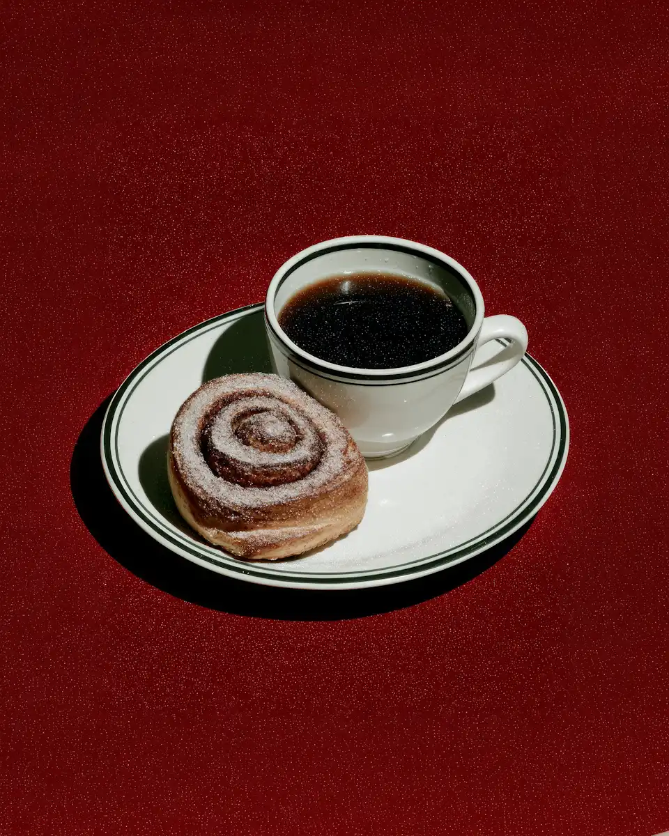 White cup of black coffee and a cinnamon roll with powdered sugar on a white plate with green stripes against a dark red background.