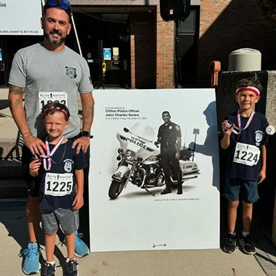 Man with two young boys wearing race numbers and medals standing by a memorial sign outdoors.