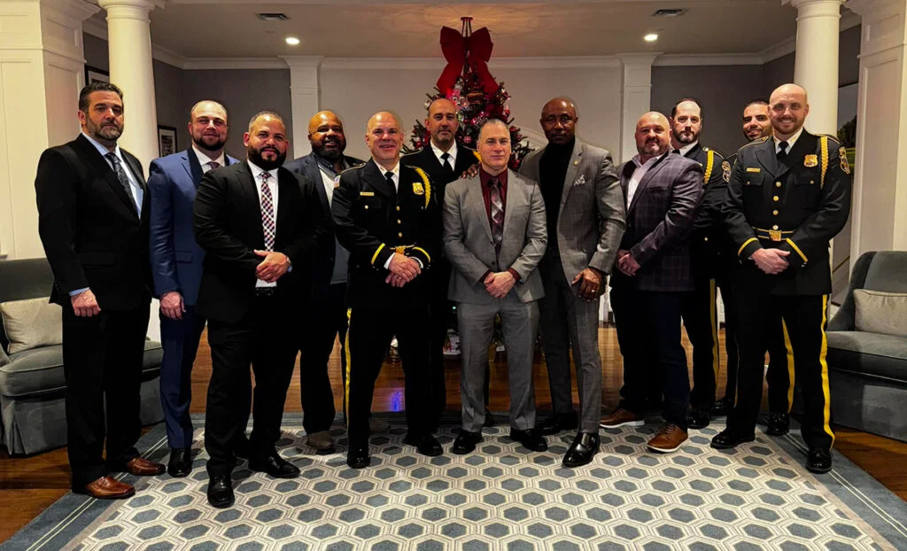 Group of men in suits and police uniforms posing indoors with Christmas tree decorated with red bow behind them.