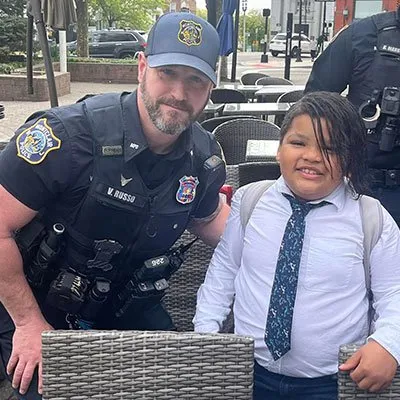 Police officer smiling and posing with a young boy dressed in a white shirt and tie at outdoor seating.