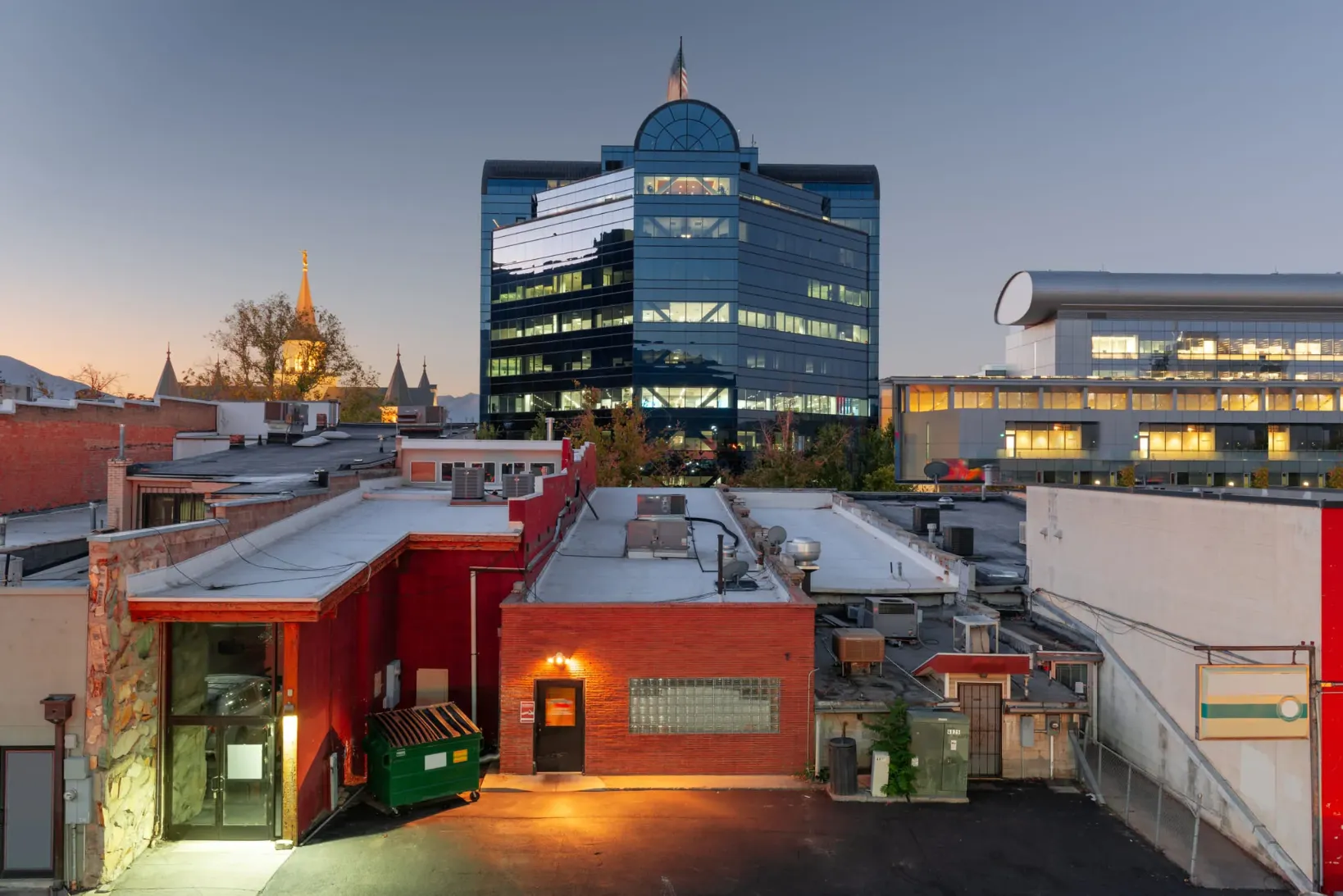 Urban cityscape at dusk with modern glass office building, illuminated older church steeple, and low-rise red brick buildings in foreground.