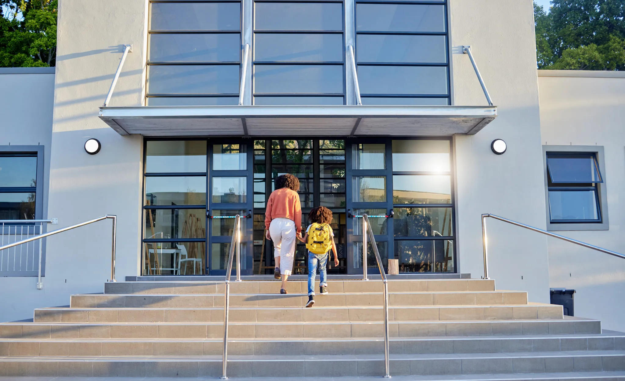 Mother and child holding hands while walking up stairs to enter a modern school building with large glass windows.