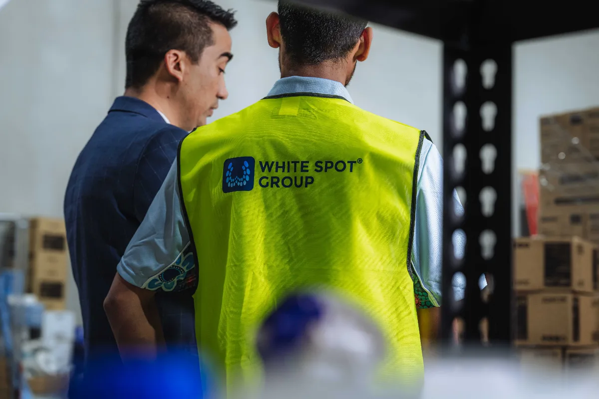 Two men in a warehouse, one wearing a bright yellow vest with 'WHITE SPOT GROUP' logo on the back.