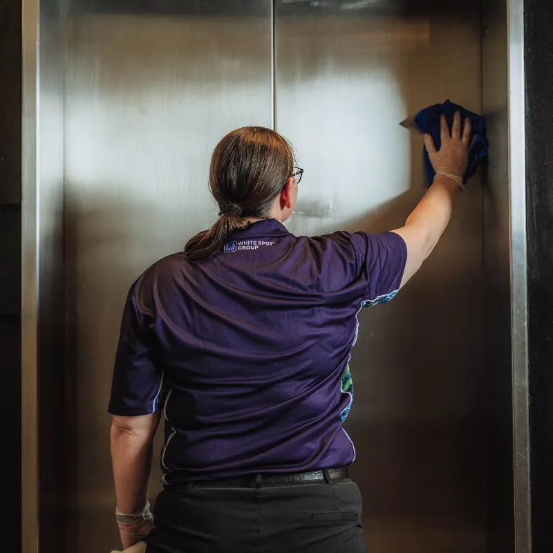 Person with ponytail wearing a Purple White Spot Group shirt cleaning a stainless steel elevator door with a blue cloth.