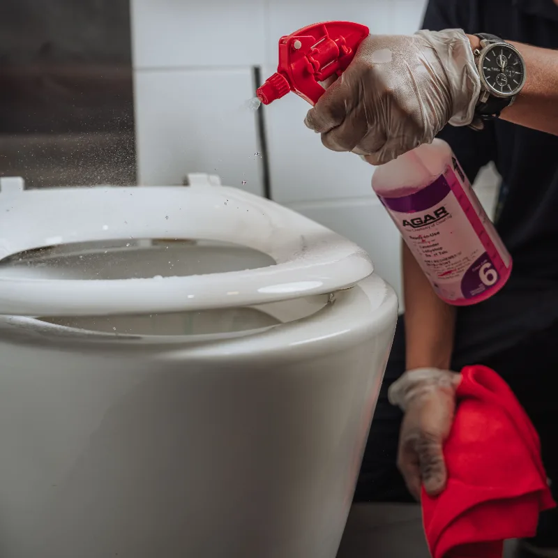 Person wearing gloves spraying disinfectant cleaner on a white toilet seat with a red cloth in hand.