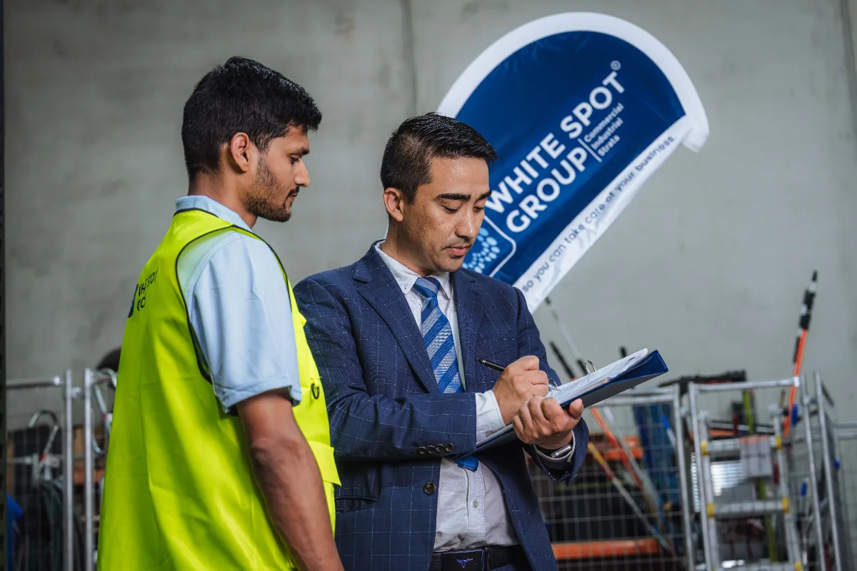 Businessman in a suit writing on a clipboard while standing next to a worker in a yellow safety vest inside a warehouse with White Spot Group banner in the background.