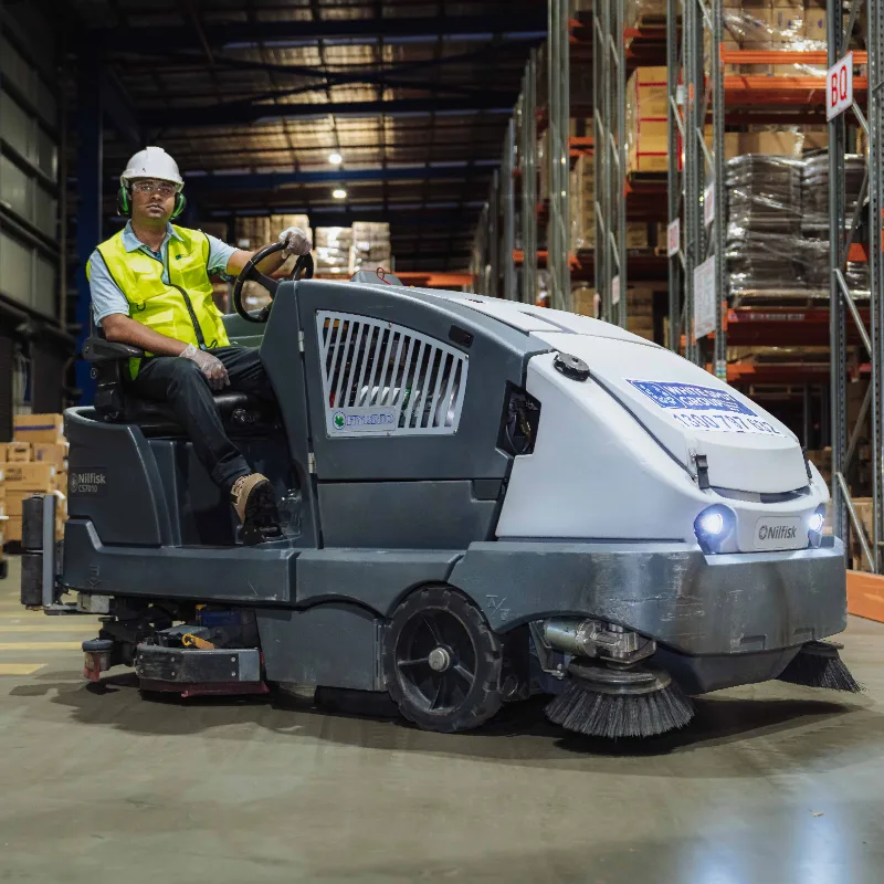 Worker wearing safety gear operating a large industrial floor cleaning machine inside a warehouse.